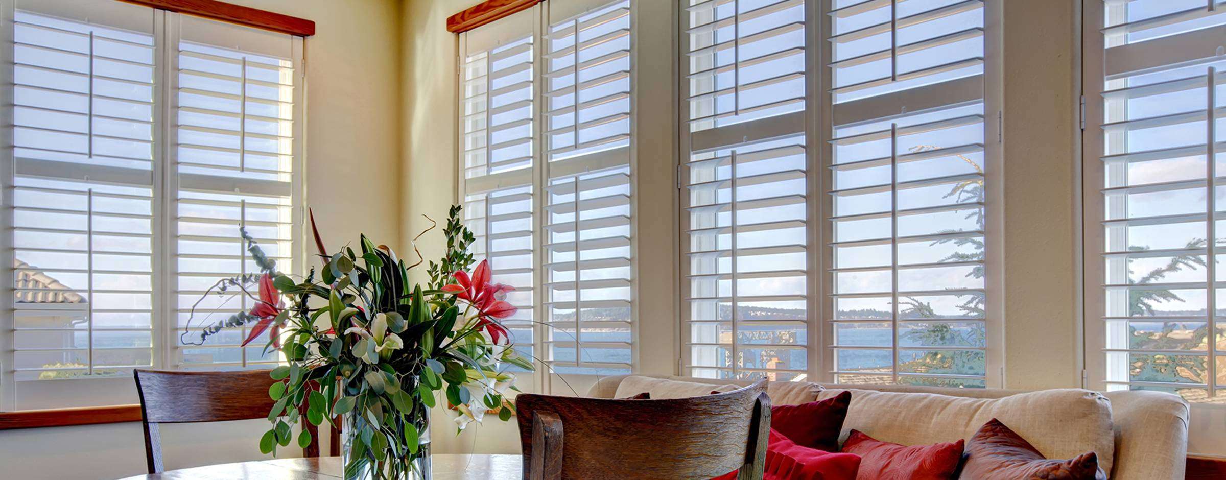 A bright living room with large white plantation shutters on the windows overlooking the water, a sofa with red pillows, and a dining table with a floral centerpiece.